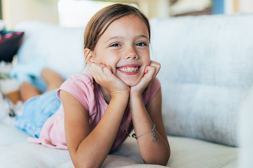 Portrait of cute little girl lying down on the sofa at home with hands on chin. Beautiful girl looking at camera and smiling relaxing at home