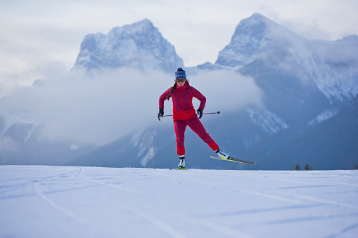 A female cross-country skier goes down a hill at the Canmore Nordic Centre Provincial Park in Alberta, Canada. She is doing the skate skiing, or freestyle, technique. Two of the Three Sisters mountains are visible in the background.