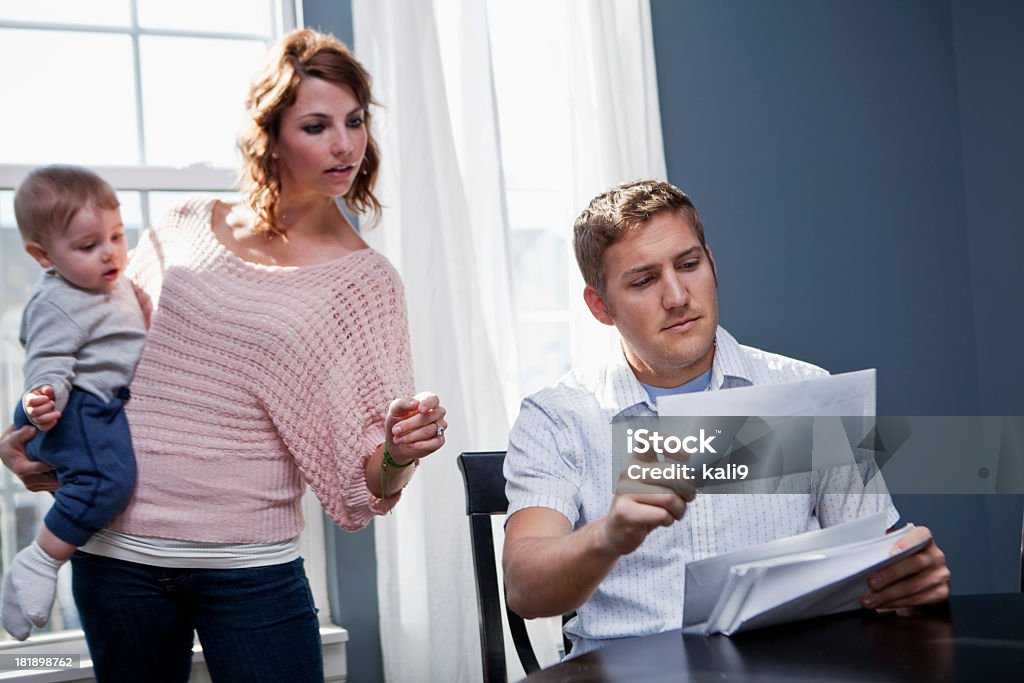 Family finances Young family at home.  Mother (20s) holding baby (8 months) while father looks through mail, paying bills.  Focus on man. Family Stock Photo
