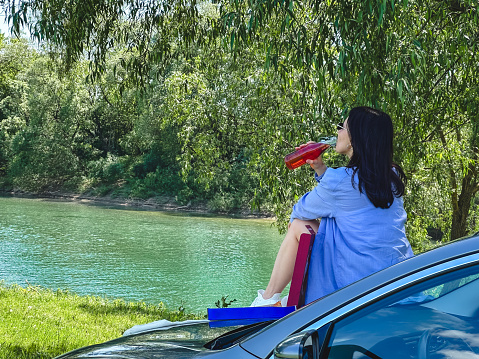 happy woman sitting on car hood eating pizza looking on river sunny summer day