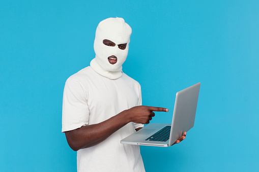 african american male hacker in white balaclava uses laptop on blue isolated background, bully in mask is typing online and pointing with his hand at the screen