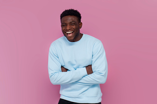 cheerful african american man in blue sweater laughing on pink isolated background, man jokes and smiles with his arms crossed