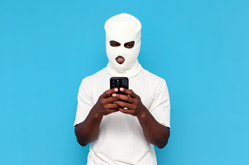 african american male hacker in white balaclava uses smartphone on blue isolated background, bully in mask is engaged in online crime on the phone