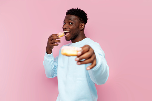 african american man in blue sweater eats sweet donuts and offers you on pink isolated background, man with pastries and food smiles and gives