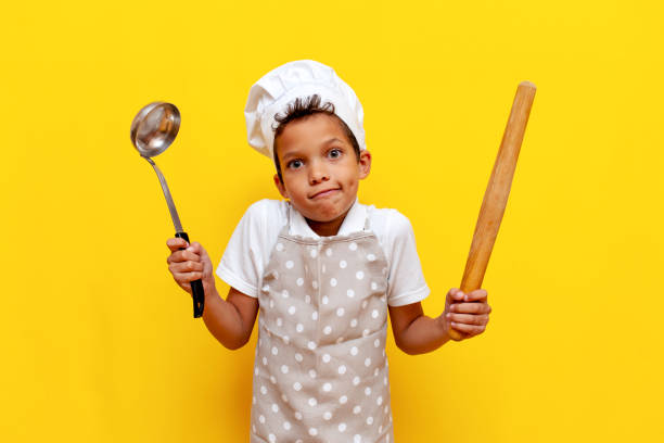 istock unsure african american boy in uniform and chef's hat holding kitchen items and shrugging on yellow isolated background 1819083268