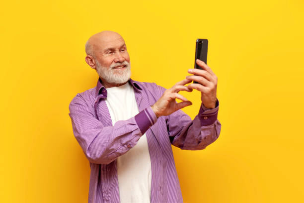 istock old bald grandfather with gray beard uses smartphone and selects online on yellow isolated background 1819083309