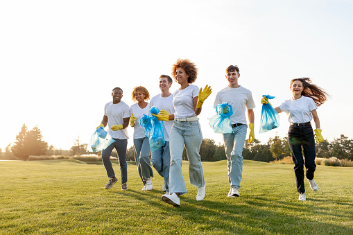 multiracial group of people running to clean the park from garbage and plastic with gloves and garbage bags, team of volunteers rejoice and celebrate outdoors, environmental care concept