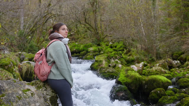 SLO MO Side View of Female Hiker Standing Near Stream Flowing Through Moss Covered Rocks in Forest