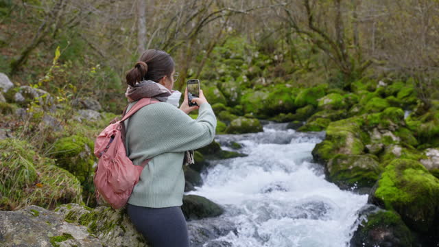 SLO MO Young Female Backpacker With Smartphone Photographing River Flowing Through Moss Covered Rocks in Forest