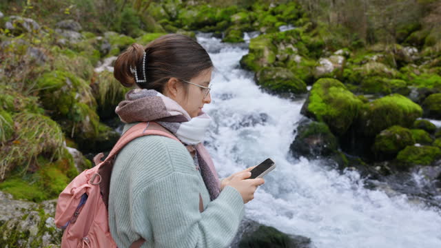 SLO MO Happy Young Female Backpacker Using Smartphone Near River Flowing Through Moss Covered Rocks in Forest