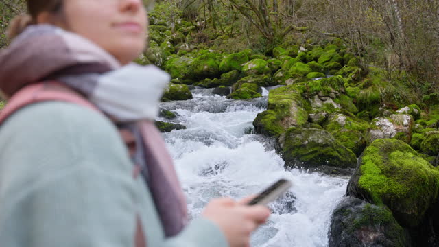 SLO MO Happy Young Woman Using Smartphone Near Focused River Flowing Through Moss Covered Rocks in Forest