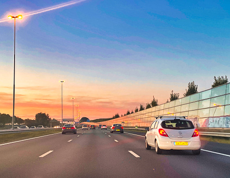 Zevenbergschenhoek, Netherlands - October 24 2021 : cars are driving on the asphalt lanes of a highway with modern transparent noise barriers in the sunset