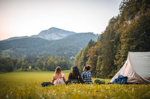 Wide angle shot of three female campers sitting on a meadow next to a tent and enjoying the amazing view. Back view of all.