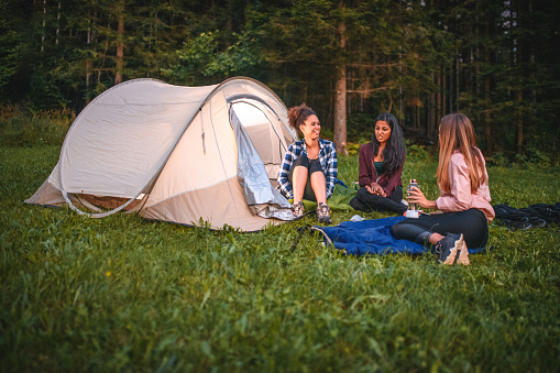 Young female backpackers talking while camping in the hills. Their tent lit from inside, all sitting in front. Dim light.