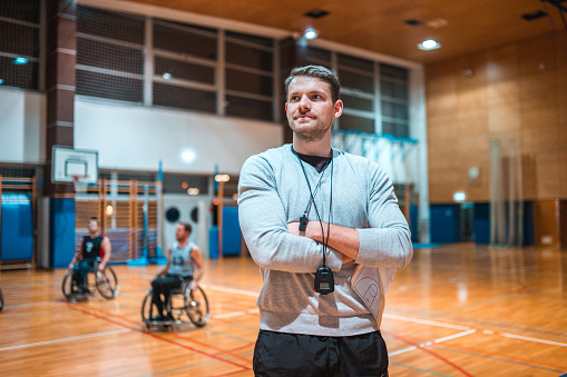 Portrait of a wheelchair basketball coach standing with his arms crossed and looking at camera while his team is practicing in the background.