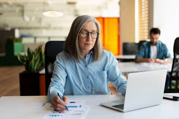 istock Mature businesswoman using laptop computer, taking notes, working project in modern office. 60 years old female, financier working with documents at workplace. Successful business, career 1819384602