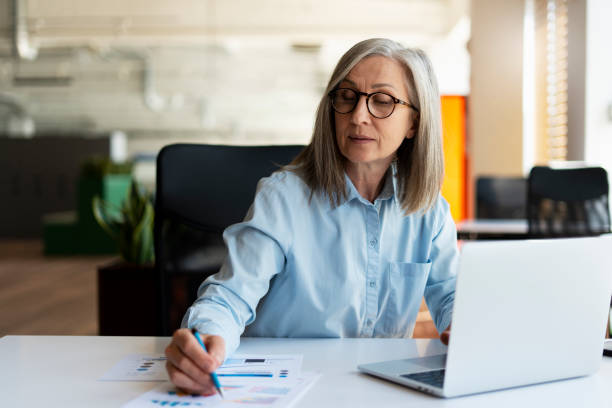 Serious mature businesswoman using laptop computer, taking notes, working project in modern office. 60 years old female, financier working with documents at workplace. Successful business