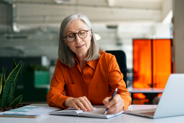 Mature woman, writer using laptop computer, taking notes, working project in modern office. 60 years old female, financier working at workplace. Successful business