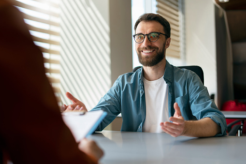 Confident handsome smiling man wearing eyeglasses sitting at job interview. Business people talking, meeting, sharing ideas in modern office. Human resources concept