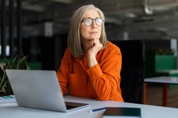 Pensive senior manager, successful gray haired businesswoman using laptop computer, working online, planning project in modern office. Serious employee looking away sitting at workplace