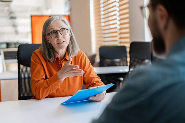 Senior business woman, employer holding resume listening young candidate at job interview. Group of business people talking, meeting, sharing ideas in modern office. Human resources concept mujer de negocios, empleador que sostiene el currículum vitae que escucha al joven candidato en la entrevista de trabajo. grupo de personas de negocios que hablan, se reúnen, comparten ideas en una oficina moderna. concepto de recursos humanos