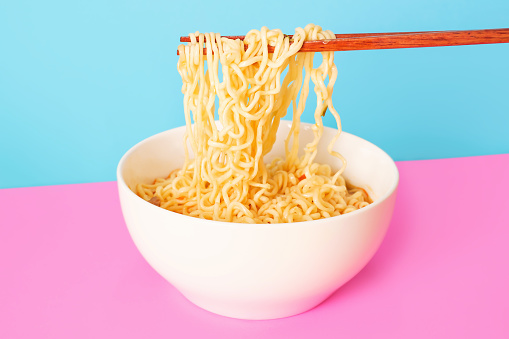 Bowl with instant noodles and chopsticks on a pink and blue background, front view.