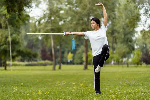 Martial arts woman practicing wushu kungfu with sword at green park. Healthy wellness active training concept