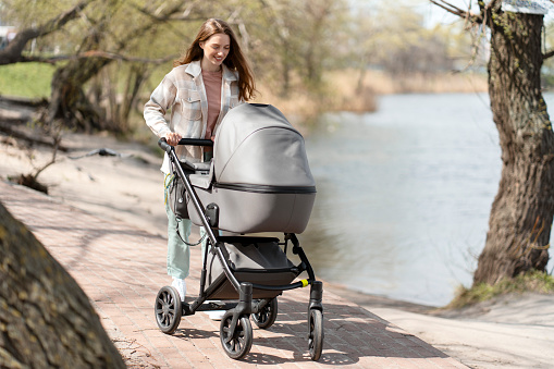 Modern, young mother walking pushing stroller with baby outdoors, walking in park. Happy woman, sunny day, cheerful motherhood