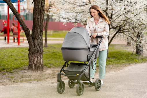 Smiling, positive woman pushing stroller with baby, walking in park, spring, sunny day. Ð¡oncept of healthy lifestyle, taking child care