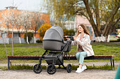 Beautiful, young mother sitting on bench, looking at stroller with baby, walking outdoors