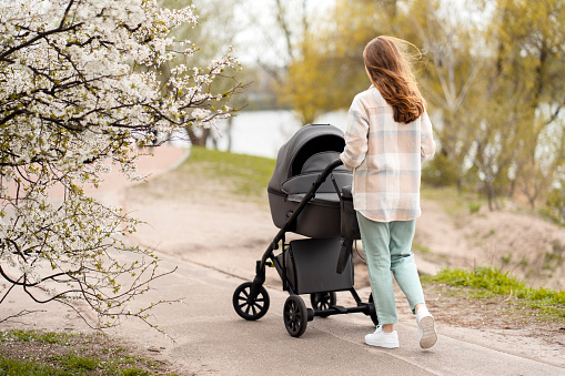 Back view, young woman walking with small child, pushing stroller, spring, sunny day. Mother and child together in nature