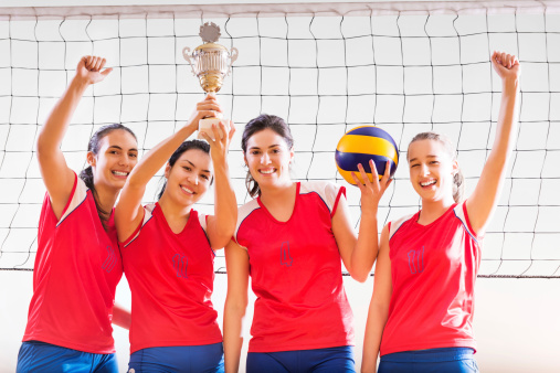Winning female volleyball team standing against the net with the champion cup raised above their heads.