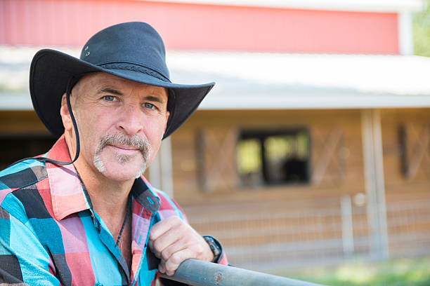 Rancher on the Farm "This is a horizontal color  photograph of a rancher on his farm. He wears plaid, jeans and a black cowboy hat. He leans on a cattle gate while looking at the camera. Depth of field is shallow. A red barn is in the background." plant city photos stock pictures, royalty-free photos & images