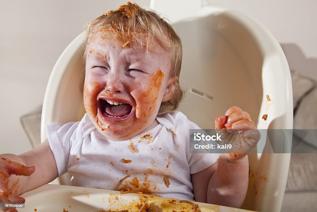 Messy baby eating, crying Messy baby (16 months) eating spaghetti in high chair, crying. Baby - Human Age Stock Photo