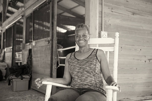 Senior Woman Sitting in a Stable "This is a horizontal, sepia toned photograph of a mature smiling African American woman. She sits in a wooden rocking chair in front of a horse stable. Depth of field is shallow." plant city photos stock pictures, royalty-free photos & images