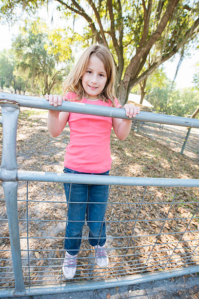 Little Girl Stands on Fence "This is a vertical color photograph of an 8 year old little girl on a farm in Plant City, Florida. She smiles and looks towards the camera while she stands on a metal fence. There is a tree in the background." plant city photos stock pictures, royalty-free photos & images
