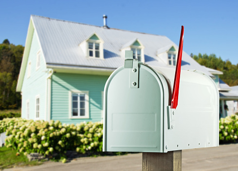 A  white country and rural mailbox with the flag raised and copy space on the mailbox In the background is a farmhouse. Selective focus on the mailbox.