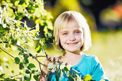 A beautiful little blonde girl smiles happily as she stands by a tree in the hazy spring sunshine.