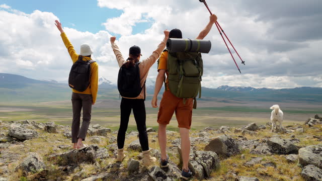 Three hikers raise arms, joy achieving success standing on top of high mountain