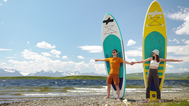 Two happy young hikers together stand with surfing boards on shore mountain lake