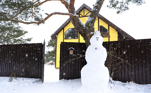Funny snowman in a hat and scarf on the background of a yellow house in the yard. Winter, winter entertainment, snowfall