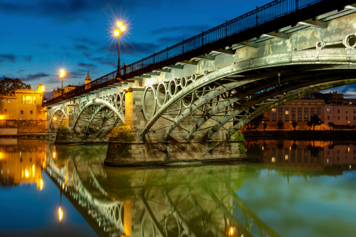The iconic Puente de Triana (Triana Bridge) crossing the Guadalquivir river in Sevilla, Andalusia, Spain. Captured at dusk with a long exposure.