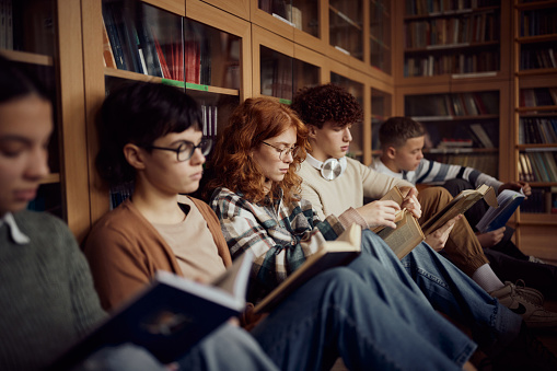 Group of high school students studying from books while relaxing on floor in library. Focus is on redhead girl.