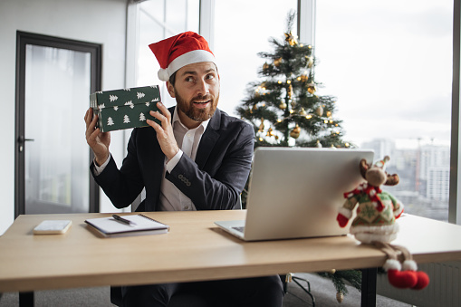Caucasian bearded businessman using laptop for live broadcasting at office on background of Christmas tree. Friendly male influencer in Santa hat sitting at desk and holding parcel box near ear.