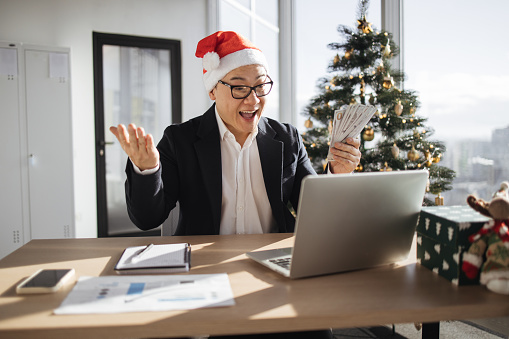 Accounting, taxes and finances concept - young excited asian attractive man in Santa hat lifting hands with cash papers counting money at modern office with Christmas tree.