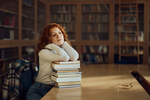 Teenage female student day dreaming while leaning on a stack of books in library.