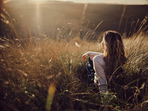 Back view of a woman looking at hill view while relaxing in tall grass at sunset. Photographed in medium format. Copy space.