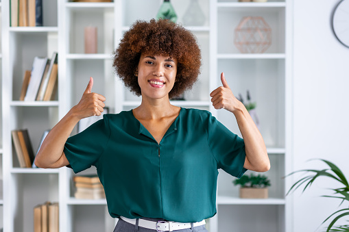 confident afroamerican woman showing two thumb up in approval, recommending, standing over home office background