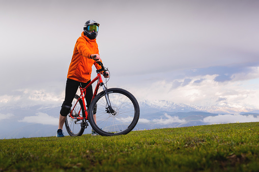 Portrait of a young cyclist standing against the backdrop of a summer landscape with a mountain in the clouds. A cyclist on a mountain path stops to watch the scenery of peaks in the distance.