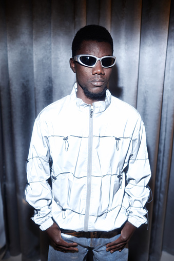 Vertical fashion portrait of young Black man wearing sunglasses and futuristic holo outfit